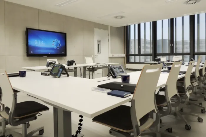 Une salle de classe grande et lumineuse au centre de formation Testia de Brême. Cinq tables avec des sièges pour les stagiaires se trouvent au centre de la pièce, face à un tableau intelligent accroché au mur derrière le bureau du formateur. Le mur de droite est entièrement recouvert par une grande façade vitrée.